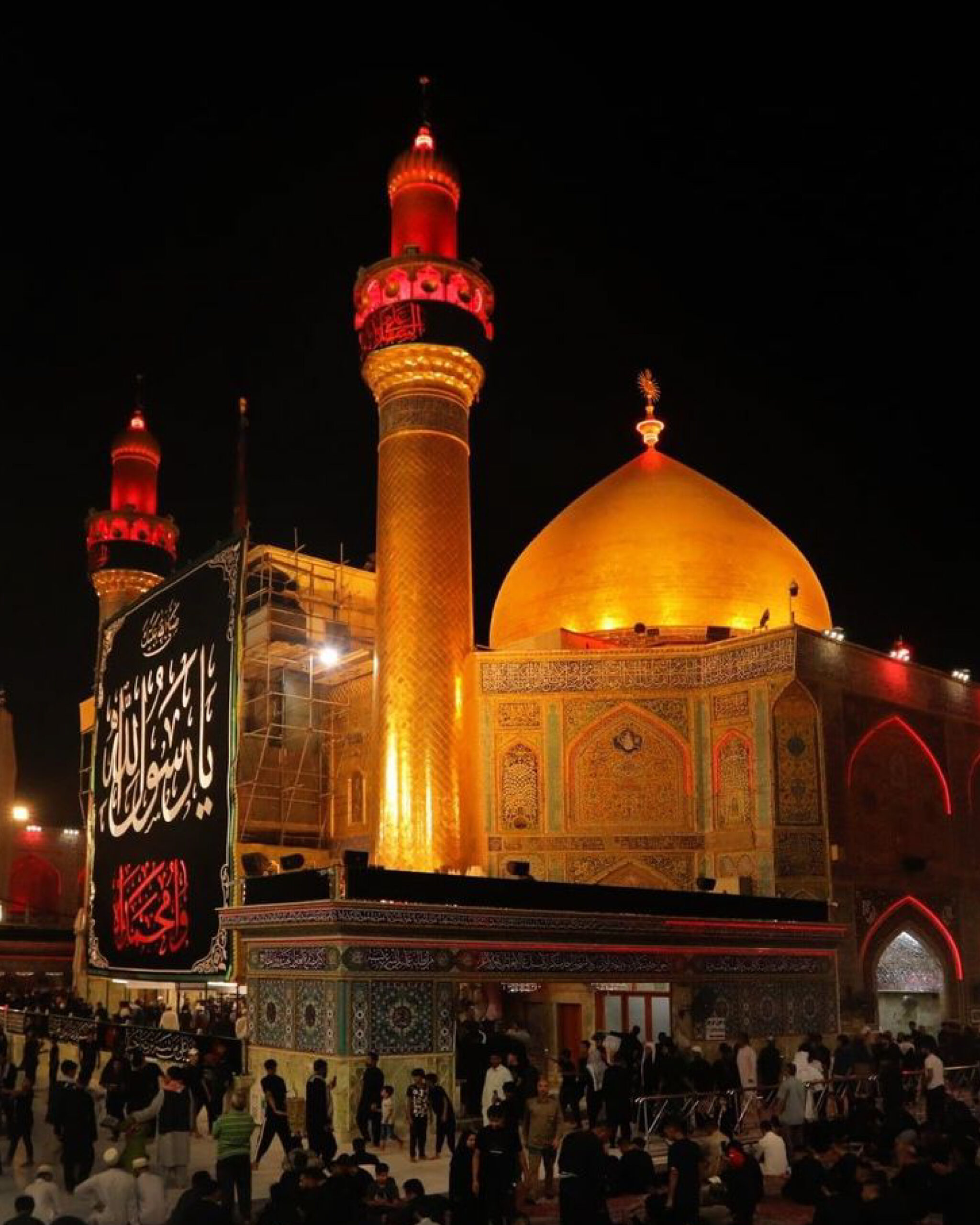 Night view of the Imam Hussain Shrine in Karbala, Iraq, with the golden dome and minarets glowing and thousands of pilgrims gathered for prayers and mourning.